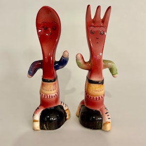 Puede incluir: Un juego de salero y pimentero de cerámica roja con forma de cuchara y tenedor con caras pintadas y ropa colorida. La cuchara lleva una camisa azul y morada y el tenedor lleva una camisa verde y amarilla.