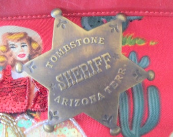 Puede incluir: Una placa de estrella de sheriff de color dorado con el texto "Tombstone Arizona Terr. Sheriff" sobre un fondo rojo con un cactus y una mujer con un sombrero de vaquero.