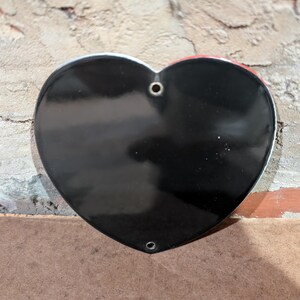 Puede incluir: Un objeto en forma de coraz&oacute;n negro brillante con un borde plateado. El coraz&oacute;n tiene dos agujeros en la parte superior y uno en la inferior. El fondo es una pared de ladrillo texturizada y una superficie marr&oacute;n.