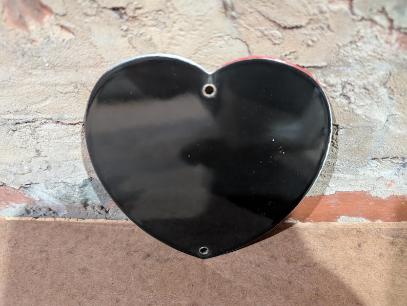 Puede incluir: Un objeto en forma de coraz&oacute;n negro brillante con un borde plateado. El coraz&oacute;n tiene dos agujeros en la parte superior y uno en la inferior. El fondo es una pared de ladrillo texturizada y una superficie marr&oacute;n.