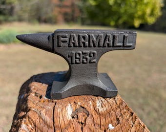May include: A dark gray cast iron anvil featuring the raised text FARMALL and the year 1952 embossed on the side. The anvil has a pointed horn on the left side and a wide, flat base, showcasing a textured, weathered metal surface with clear lettering.