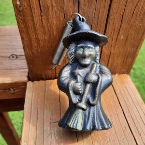 May include: A black cast iron witch figurine with a green hat and green eyes. The witch is holding a broom in her right hand. The figurine is attached to a spring.