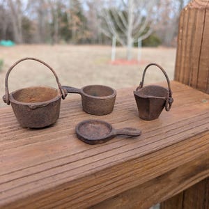 May include: Four small, rustic, brown metal containers sit on a wooden surface. There is a small bucket with a handle, a small scoop, a small, faceted bucket with a handle, and a small, round scoop.