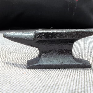 May include: A small, black anvil with a flat top and a pointed horn. The anvil has a rectangular base and a curved body. It is made of metal and appears to be a miniature decorative item.
