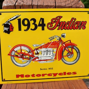 May include: A yellow and red vintage metal sign advertising 1934 Indian Motorcycles, Series 402. The sign features a red motorcycle with a chrome engine and white wheels. The sign also features a red and white illustration of an Indian head with a feather headdress.