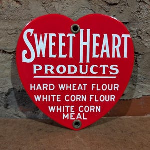 Puede incluir: Un letrero rojo en forma de coraz&oacute;n con las palabras blancas "SWEET HEART PRODUCTS", y la lista "HARD WHEAT FLOUR", "WHITE CORN FLOUR" y "WHITE CORN MEAL". El letrero tiene dos agujeros.