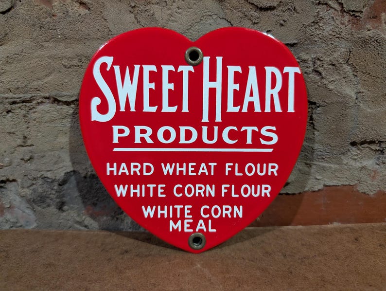 Puede incluir: Un letrero rojo en forma de coraz&oacute;n con las palabras blancas "SWEET HEART PRODUCTS", y la lista "HARD WHEAT FLOUR", "WHITE CORN FLOUR" y "WHITE CORN MEAL". El letrero tiene dos agujeros.