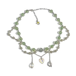 May include: A delicate silver necklace with a double strand of green and white iridescent beads. The necklace features a heart-shaped pendant and three dangling crystal beads.