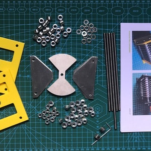 Könnte beinhalten: Ein Satz Teile zum Zusammenbauen eines Schmetterlingskondensators, einschließlich Metallplatten, Stäbe, Muttern und Lagern. Die Teile liegen auf einer grünen Schneidematte mit einem gedruckten Raster. Das Bild enthält auch Anweisungen zum Zusammenbauen des Kondensators.