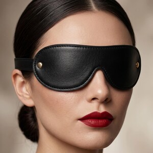 Könnte beinhalten: Eine schwarze Leder-Augenbinde mit geschwungenem Design und Riemen. Die Augenbinde hat goldfarbene Nieten auf jeder Seite. Die Person im Bild hat dunkles Haar und roten Lippenstift.