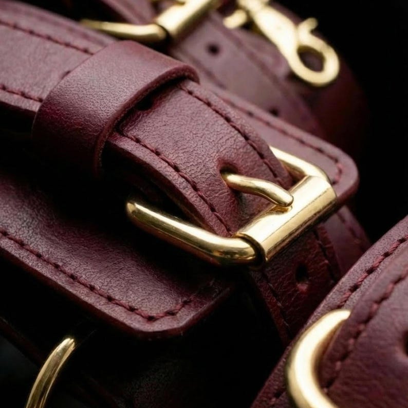 Close-up of luxury burgundy leather cuffs with gold hardware, premium handmade BDSM accessories for couples and romantic bedroom play.