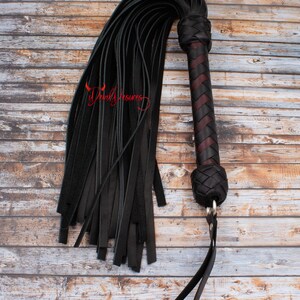 May include: A black leather whip with a braided handle and a long, fringed tail. The handle is made of dark brown leather with a red accent.