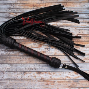 May include: A black leather whip with a braided handle and a red accent. The whip is lying on a wooden surface.