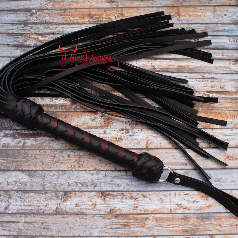 May include: A black leather whip with a braided handle and a red accent. The whip is lying on a wooden surface.