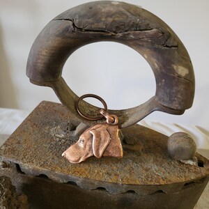May include: A copper dog pendant with a keyring hangs in front of a vintage iron. The pendant is a detailed profile of a dog's head. The iron has a wooden handle and a rusty, aged appearance, suggesting it is antique.