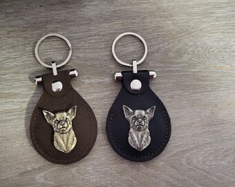 May include: Two leather keychains with metal key rings. Each keychain features a silver-colored Chihuahua dog head charm. One keychain is brown leather and the other is black leather.