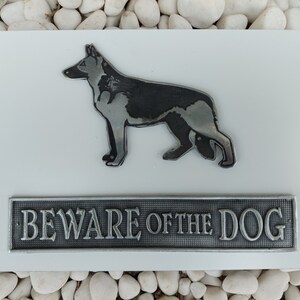 Könnte beinhalten: Ein weißes Metallschild mit einer schwarzen und silbernen Silhouette eines Deutschen Schäferhundes und dem Text "BEWARE OF THE DOG".