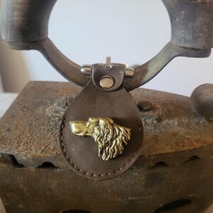 May include: A brown leather dog tag with a gold-colored dog head emblem. The tag is attached to a metal clasp. The background includes a vintage iron and a wooden handle.