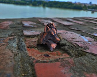 May include: A copper-colored wolf head pendant with intricate details. The pendant is sitting on a brick surface.