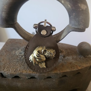 Könnte beinhalten: Ein brauner Leder-Schlüsselanhänger mit einem metallischen Hundeemblem. Der Hund ist eine detaillierte, goldfarbene Skulptur. Der Schlüsselanhänger hat einen silberfarbenen Verschluss und eine kleine, runde silberne Niete. Das Leder ist oval und hat einen genähten Rand.
