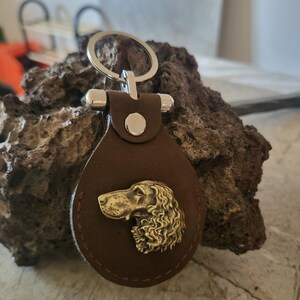 May include: A brown leather keychain with a metal dog emblem. The keychain has a silver ring and accents. The dog emblem is a detailed, golden-colored relief of a dog's head.