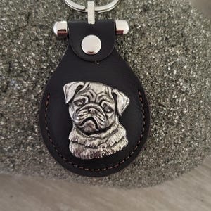 May include: A black leather keychain with a silver pug charm. The keychain has a silver ring and a silver button closure.