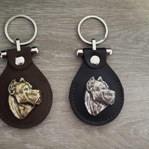 May include: Two leather keychains with metal key rings. Each keychain features a silver-colored dog head silhouette. One keychain is brown leather and the other is black leather.