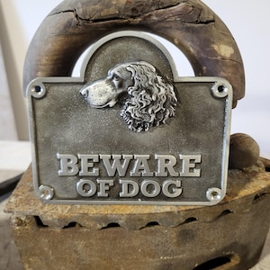Könnte beinhalten: Rechteckiges Metallschild mit dem erhabenen Schriftzug „BEWARE OF DOG“. Das Schild zeigt ein detailliertes Relief eines Hundekopfes und ist auf einem rustikalen Holzsockel montiert. Das Schild ist eine Warnung.