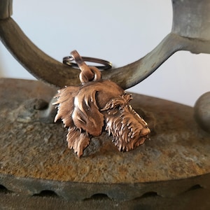 Pode incluir: Um porta-chaves de cabeça de cão cor de cobre. O pendente apresenta detalhes de pêlo e traços faciais, preso a um pequeno anel de metal. O porta-chaves é exibido contra um fundo escuro e texturizado.