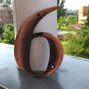 Puede incluir: Primer plano de un número de casa seis de metal de color cobre. El número está sobre una superficie gris con un fondo borroso de árboles y edificios.