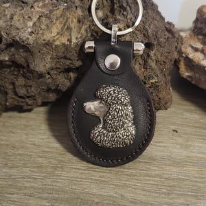 May include: A black leather keychain with a silver poodle charm. The keychain has a silver ring for attaching keys.