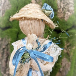 May include: A straw doll wearing a straw hat with a blue and green ribbon. The doll is holding a bouquet of dried flowers and a blue ribbon.