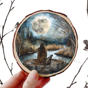 May include: A round wooden slice with a painted scene of a woman and a beaver sitting by a river under a full moon. The scene is painted in shades of blue, green, and brown.