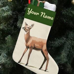 Puede incluir: Un calcetín navideño con una ilustración de un ciervo. El calcetín tiene un cuerpo color crema con un ciervo marrón y un puño verde con el texto "Your Name". Está colgado en un árbol de Navidad.