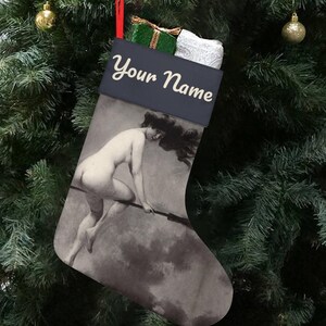 Puede incluir: Un calcetín navideño con una parte superior negra y una imagen en tonos sepia de una mujer montada en una escoba. El texto "Your Name" está impreso en escritura blanca. El calcetín está colgado de un árbol de Navidad con adornos.