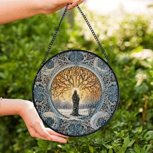 May include: Round stained glass panel featuring a tree with a figure in a winter landscape. The artwork is framed by a decorative border and suspended by a black chain. The color palette includes shades of blue, gold, and white.