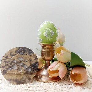 May include: A green Easter egg with a white floral design sits on a gold metal stand. The egg is surrounded by pastel pink tulips.