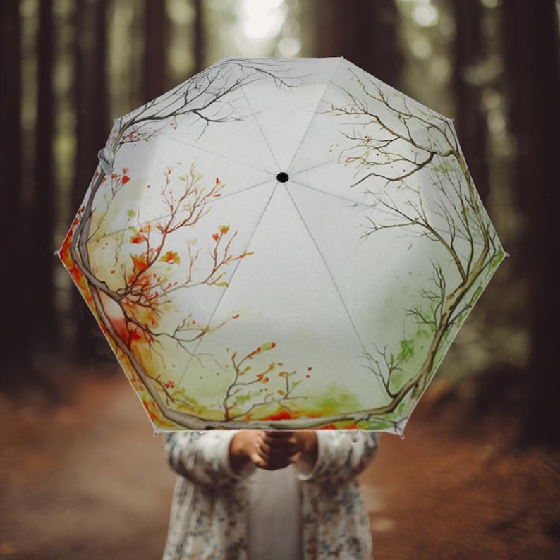 K&ouml;nnte beinhalten: Ein wei&szlig;er Regenschirm mit einem Aquarelldesign von &Auml;sten und Herbstbl&auml;ttern in Rot-, Orange- und Gr&uuml;nt&ouml;nen. Der Schirm ist ge&ouml;ffnet und wird vor einem verschwommenen Waldbild gehalten.