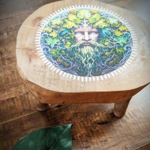 Peut inclure: Un petit tabouret rond en bois à trois pieds. Le dessus présente une illustration colorée de l'Homme Vert, un visage entouré de feuilles et de feuillage. Le tabouret est en bois naturel avec un grain visible.