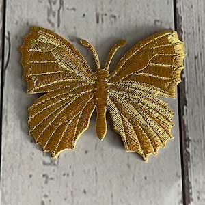 Può includere: Un'applicazione di farfalla ricamata in oro. La farfalla ha un design dettagliato con una finitura testurizzata e metallica.