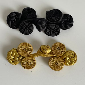 Puede incluir: Dos juegos de cierres de rana decorativos. Un juego es negro y el otro dorado. Cada cierre presenta una serie de nudos en espiral y un lazo de conexión, diseñados para sujetar prendas u otros artículos de tela. Sobre un fondo blanco.