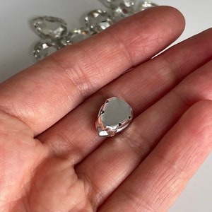 May include: A silver-colored, teardrop-shaped rhinestone with a flat back and a faceted design.