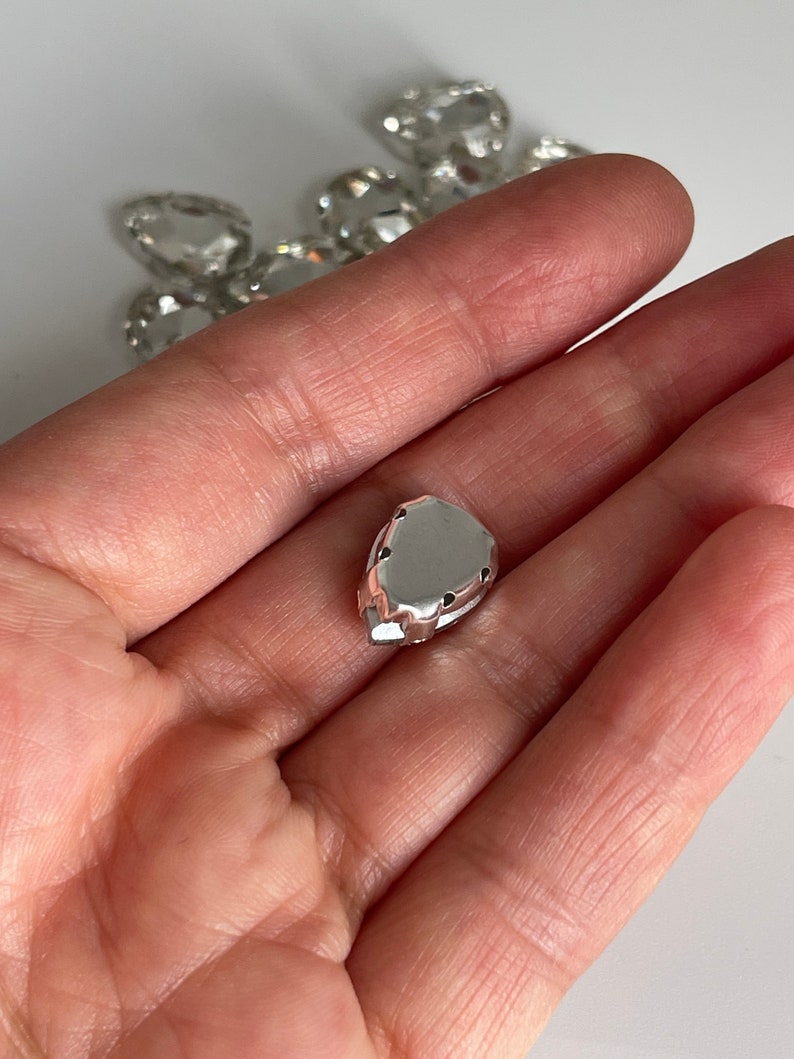 May include: A silver-colored, teardrop-shaped rhinestone with a flat back and a faceted design.