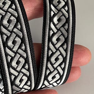 Può includere: Nastro decorativo nero e argento con un motivo a nodo celtico ripetuto. Il nastro ha un bordo nero e un motivo argentato intrecciato dettagliato. Adatto per artigianato, cucito o abbellimenti.