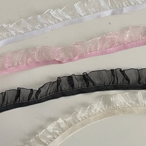 Könnte beinhalten: Vier verschiedene Arten von Rüschenbesatz mit elastischem Rücken. Die Besätze sind weiß, rosa, schwarz und elfenbein.