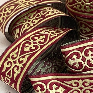 Puede incluir: Primer plano de rollos de cinta decorativa. La cinta presenta un fondo burdeos con un diseño bordado dorado y ornamentado. La cinta está enrollada y apilada, mostrando el intrincado patrón y la textura. Ideal para manualidades y adornos.