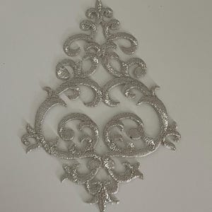 May include: Silver embroidered applique with a symmetrical, ornate design. The applique features swirling patterns and a central, stylized floral element. The embroidery has a textured appearance, creating a dimensional effect. The applique is likely for embellishing clothing or crafts.