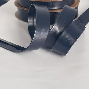 May include: Navy blue leather ribbon spools. The ribbon is a solid color and appears to be made of a smooth, durable material. Several spools are stacked, with a length of ribbon unrolled in the foreground.