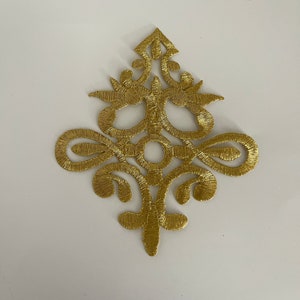Puede incluir: Aplique bordado en oro con un diseño decorativo e intrincado. El aplique tiene forma de diamante con una parte superior puntiaguda y una parte inferior redondeada.