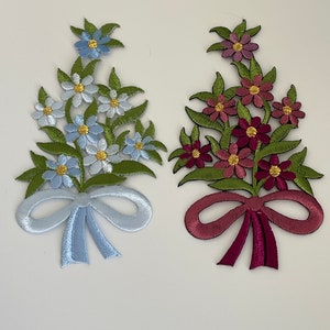 Peut inclure: Deux bouquets de fleurs brodés avec des nœuds. Le bouquet de gauche a des fleurs blanches et bleues avec un nœud bleu clair. Le bouquet de droite a des fleurs roses et rouges avec un nœud bordeaux.